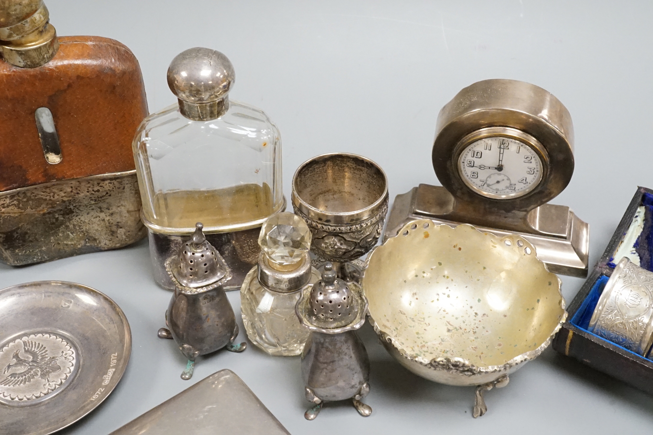 A George V silver mounted small mantel clock, height 93mm, a pair of silver pepperettes and eight other items including a silver mounted cigarette box, a cased set of four chased silver napkin rings and two plated hip fl